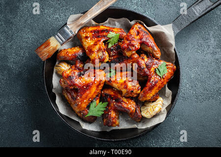 Baked chicken wings in barbecue sauce with sesame seeds and parsley in a cast iron pan on a dark concrete table. Top view. Stock Photo