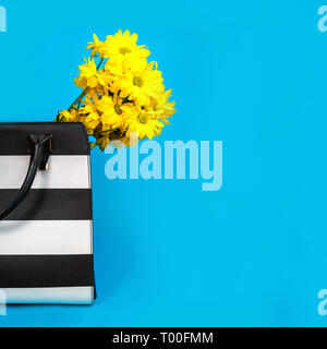 Beautiful yellow flowers and black and white trendy female bag on blue background. Stock Photo