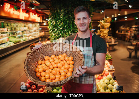 Young man stand at fruit boxes in grocery store. He hold basket with oranges and pose on camera. Positive young man smile. Stock Photo