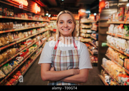 Busy young woman stand in line between shelfs with pasta. She pose on camera and smile. Happy positive model. Stock Photo