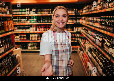 Young woman stand in alcohol shelfs in grocery store. She reaxh hand to camera and smile. Cheerful happy young woman. Stock Photo