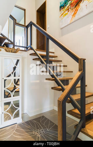 Hickory wood stairs with black metal and wood top clear glass railing leading from basement room to main floor inside luxurious contemporary home. Stock Photo