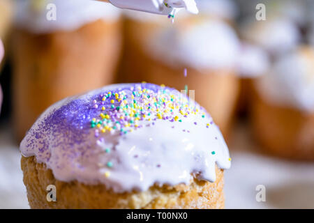 Baker decorates the upper part of Easter cakes covered with white sugar icing by colored granulated sugar pouring decoration on fresh icing making tra Stock Photo