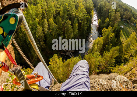 Climber's legs hanging on a rope in a harness, first person view to river in forest Stock Photo