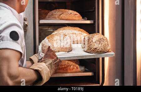 Tasty and fresh. Baker's hands in working gloves taking out freshly baked bread from the oven at the kitchen. Bakery concept. Stock Photo