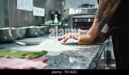 Making pasta. Cropped image of professional chef with tattoos on his hands kneading the dough in restaurant kitchen. Close up Stock Photo