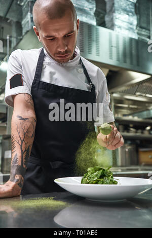 Healthy eating. Vertical portrait of handsome male chef in black apron adding spices in salad while standing in a restaurant kitchen. Stock Photo