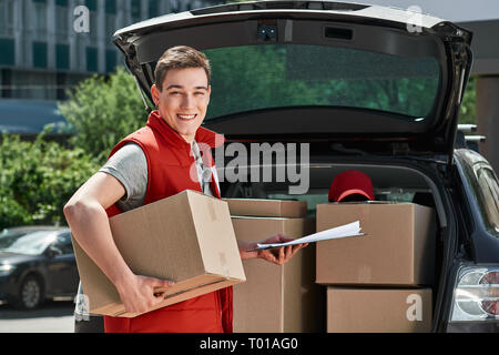 Smiling delivery man in red postal uniform standing near trunk of his car with a box and a clipboard in his hands. He is ready to do his job. Friendly worker, high quality delivery service. Outdoors. Stock Photo