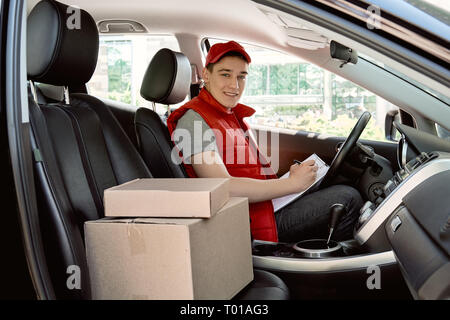 Parcel delivery man in red postal uniform drives a car. He is looking at the camera with a smile. Transport postal delivery courier delivering packages to customers. Impressive service. Stock Photo