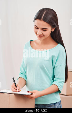 Young entrepreneur, SME, freelance woman working online, business by using smartphone, making purchase, order and preparing packages. Woman standing near stack of boxes, making notes on a clipboard. Concept of success Stock Photo
