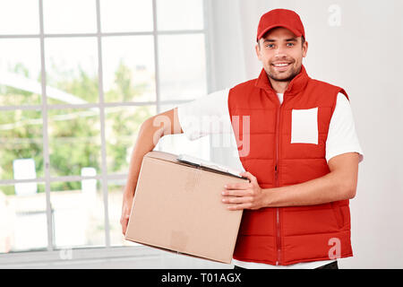 Cheerful postman wearing red postal uniform is delivering parcel to a client. He holds carton box and looking at the camera with a smile. Friendly worker, high quality delivery service. Indoors. Bright interior. Stock Photo