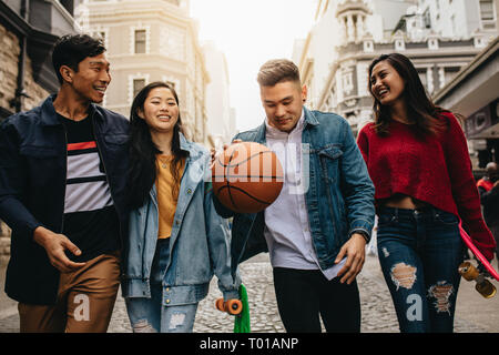 Group of friends walking together on the street, with women holding skateboards and man bouncing basketball. Stock Photo