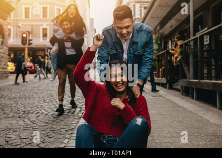 Excited woman being pushed on skateboard by man outdoors on street, with friends piggybacking in background on the city street. Group of friends hangi Stock Photo