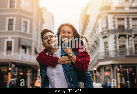 Asian man carrying his girlfriend on his back walking on the city street. Couple piggybacking on city street. Stock Photo