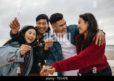 Diverse group of young people celebrating new year's day at the beach. Young asian people having fun with sparklers outdoors at the sea shore. Stock Photo