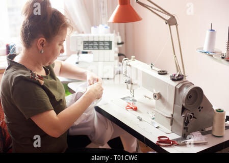 Clothier sitting in atelier, stitching white dress with hands and needle. Seamstress workplace with sewing machine, overlock, scissors, pins, threads, Stock Photo