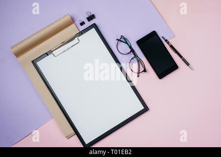Creative flat lay, top view office table desk. Workspace with blank clip board, office supplies, pen, notepad, smartphone, eyeglasses on pink and viol Stock Photo