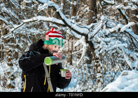 Bearded man in an elf hat in the forest pours hot tea from a thermos Stock Photo