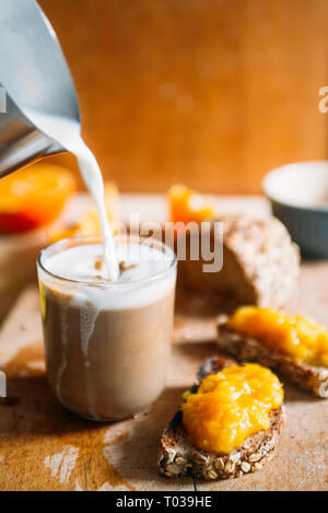 Homemade Orange jam  on a natural raw wheat bread and orange  slices while pouring milk on a  cup of coffee over a wooden cutboard, healthy breakfast, Stock Photo