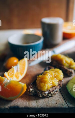 Homemade Orange jam  on a natural raw wheat bread and orange  slices and a cup of coffee and milk over a wooden cutboard, healthy breakfast,close up a Stock Photo