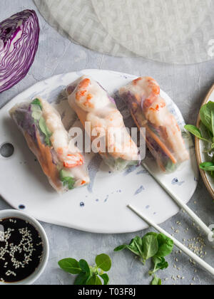 Spring rolls with carrots, red cabbage, corn lettuce and shrimps on a white ceramic board. Soy sauce with sesame seeds on a gray background Stock Photo