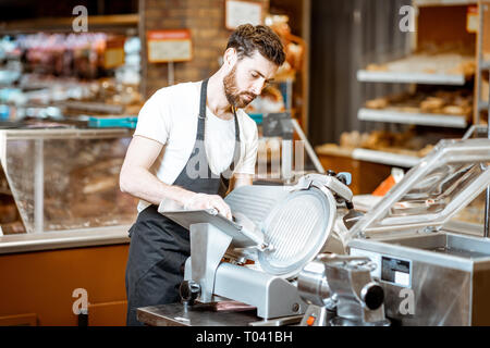 Handsome worker in uniform slicing cheese with cutting machine in the supermarket Stock Photo