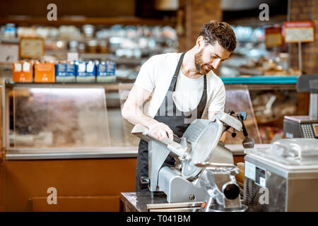 Handsome worker in uniform slicing cheese with cutting machine in the supermarket Stock Photo