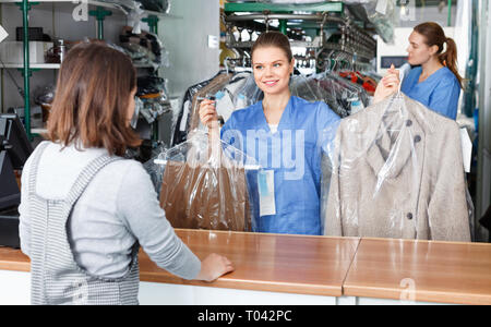 Diligent friendly smiling girl worker of laundry regretfully excusing while giving clothes after dry cleaning to female client Stock Photo