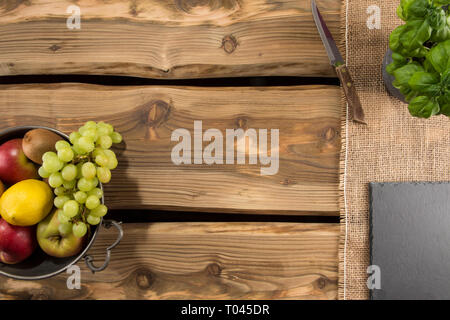 Space for text on pure wooden planks with arranged rustic elements like fruits in a zinc bowl, jute cloth, old knife, slate board. Shot from above. Stock Photo