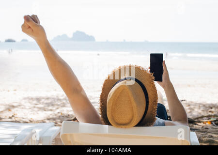 Unrecognizable guy in straw hat clenching fist and browsing smartphone while relaxing on lounger near sea Stock Photo