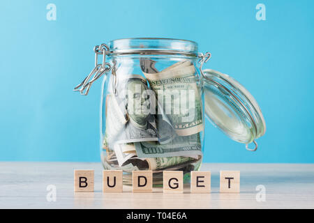 glass jar filled with dollars and word budget made of wooden blocks on blue background Stock Photo