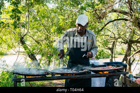 Black man cooking breakfast on outdoor grill with bacon and sausages for breakfast for Safari guests, Sabi Sands game reserve, South Africa Stock Photo