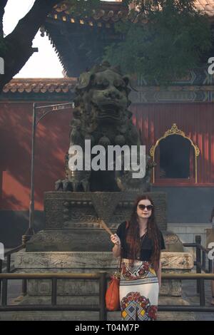 Long haired brunette lady wearing sunglasses and fashionable clothes. Holding incense in a buddhist Chinese temple, posing in front of a lion or tiger Stock Photo