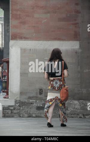 Long haired brunette hippie caucasian western female tourist walking away from camera, wearing fashionable split skirt / trousers and carrying an oran Stock Photo
