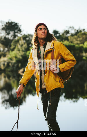 Man exploring nature walking in a forest beside a lake. Young tourist carrying a backpack walking outdoors holding a stick. Stock Photo