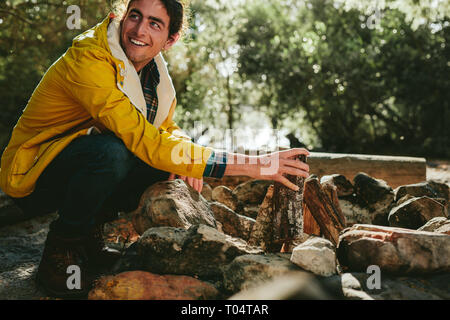 Smiling man putting fire wood setting up a bonfire in the countryside. Side view of a cheerful young man camping in a forest arranging fire wood. Stock Photo