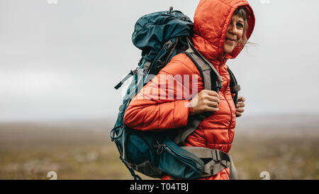 Side view of a woman hiker standing on top of a hill. Senior woman wearing a hooded jacket and a backpack standing on top of a hill during a trek. Stock Photo