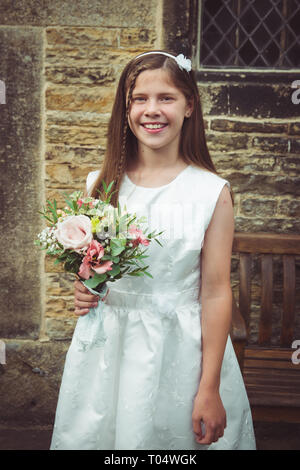 Beautiful smiling tween or teen young bridesmaid holding a rustic wildflower bouquet, wearing a white dress, in front of a vintage stone church Stock Photo