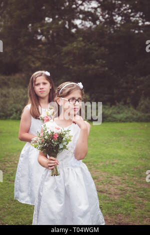 Sweet child and young teen girl bridesmaids wearing short white dresses, holding hand-tied bouquets with foliage in a park waiting Stock Photo