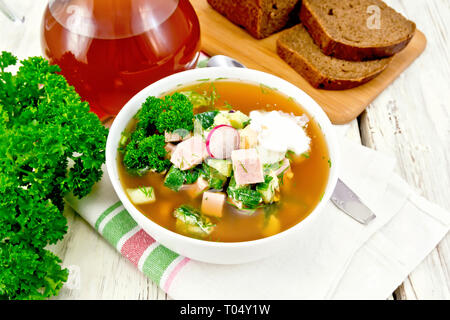 Cold soup okroshka from sausage, potatoes, eggs, radish, cucumber, greens and kvass in a white bowl on napkin, bread and jug with drink on the backgro Stock Photo