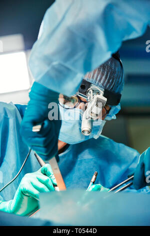 Doctor during the surgery in the operation room. She wears a blue mask, colorful hat and glasses. His assistant is next to him. Close Up. Horizontal. Stock Photo
