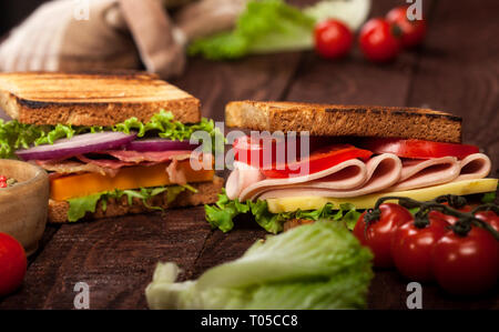 Fresh delicious toasts with cheese, tomato, onion, ham, prosciutto, cucumber and lettuce on wooden table and dark background Stock Photo