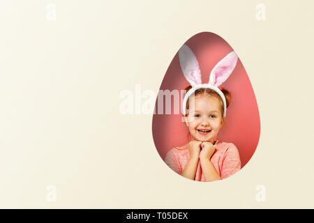 Cute little baby girl with bunny ears. A child in a hole in the shape of an egg on a colored pink and white background. Easter concept. Stock Photo