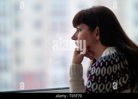 Young pretty brunette woman with long hair looks thoughtfully while standing at the window close-up, against the background of houses, against the bac Stock Photo