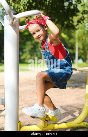 beautiful little girl in denim overalls and red kerchief, white sneakers  is playing at children's playground. kid is making steps on sports simulator Stock Photo