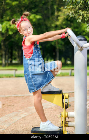 pretty little girl in denim suit and red bandana plays on children's playground. the kid is engaged on sports simulator outdoors in the park Stock Photo
