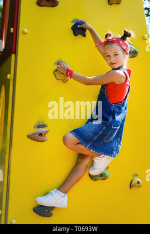 little girl with hairstyle tail in jeans jumpsuit climbs up the wall for climbing Stock Photo