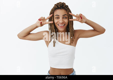 Waist-up shot of amused joyful and emotive stylish dark-skinned modern woman with dreadlocks showing peace signs with hands over eyes smiling having Stock Photo