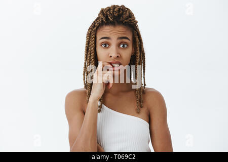 Worried and concerned silly african american attractive young woman with dreads in white top biting finger and staring guilty and nervous at camera Stock Photo