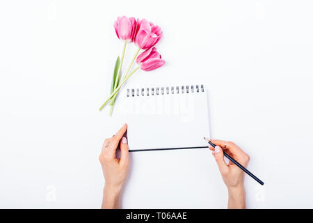 Top view female's hands writing in empty notebook next to bouquet of fresh pink tulips on white table, copy space. Stock Photo